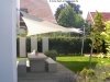 Ein weißes Sonnensegel von Lisori schützt eine Terrasse mit Tisch und Stühlen vor einem Haus. Die Befestigungen sind aus V2 Edelstahl. Ein entspannter Ort zum Verweilen!
