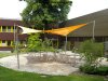 Ein großes gelbes Sonnensegel in Viereckform freistehend ausgeführt mit vier Segelmasten beschattet eine bestuhlte Terasse auf einer Außenfläche | Lisori Sonnensegel