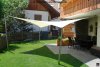 Ein weißes fünfeckiges Sonnensegel von Lisori schützt eine Terrasse mit Tisch. 3 Edelstahlmasten und 2 Aufhängepunkte an der Terasse halten das Segel.